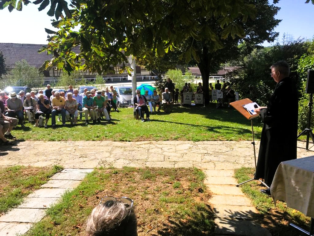 Open-Air-Gottesdienst auf einer Wiese mit Pfarrer am Pult, gedecktem Altartisch, Gemeinde auf Bänken und Bläsern im Hintergrund