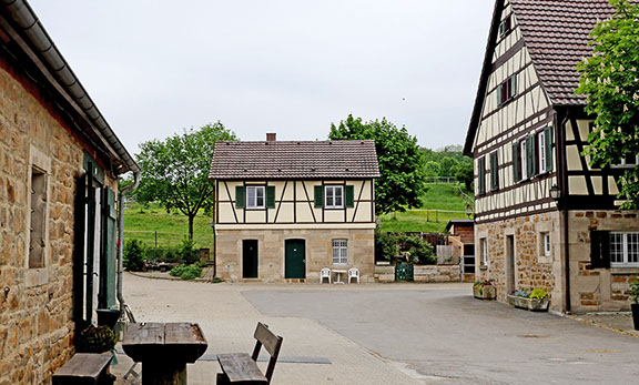 Sonniger Hof mit Sitzgruppe im Vordergrund, umrahmt von Fachwerk- und Natursteingebäuden und grüner Landschaft