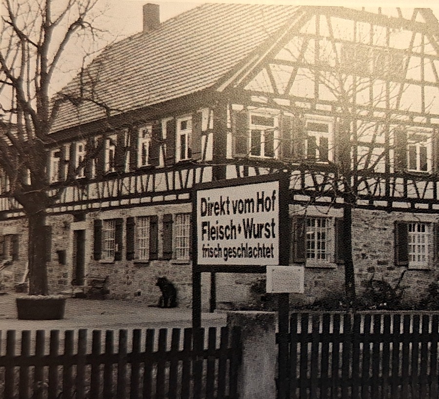 Schwarzweißaufnahme des Fachwerk-Verwalterhauses mit großem Pfostenschild Direkt vom Hof Fleisch und Wurst frisch geschlachtet, davor ein Lattenzaun