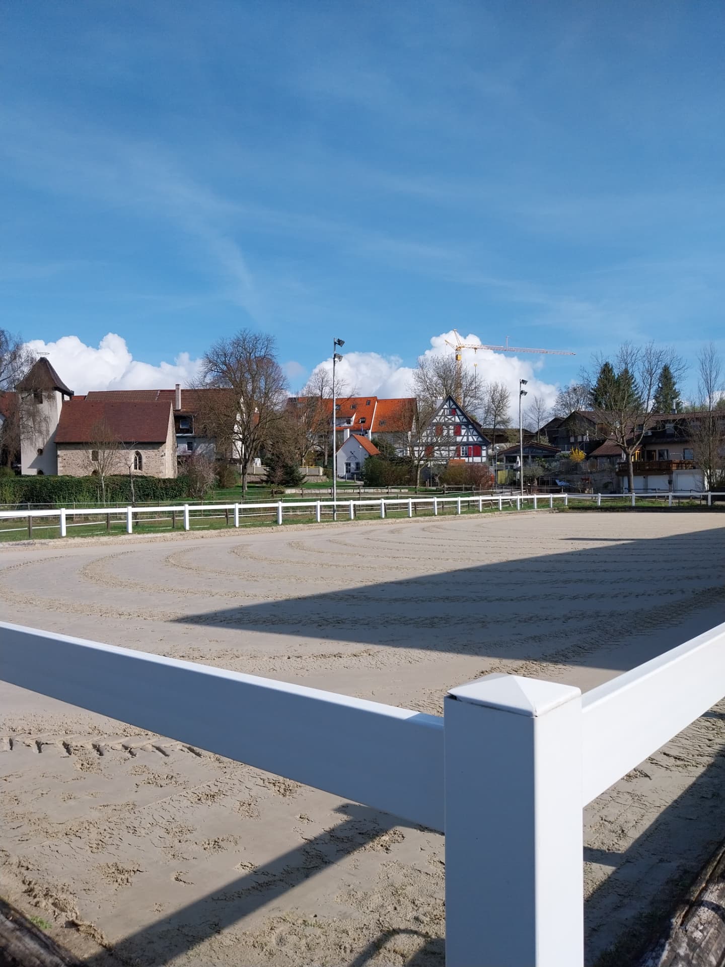 Frisch geebneter Sand-Außenplatz mit weißem Zaun, dahinter die Kapelle und Fachwerkhäuser des Dorfs