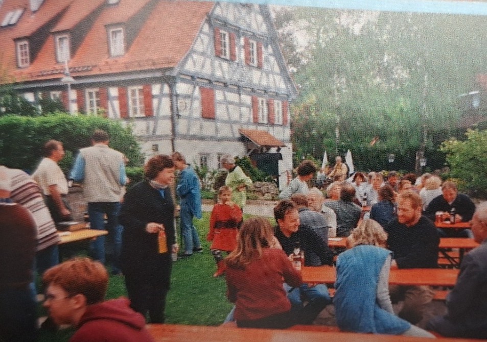 Festgesellschaft an Tischen auf dem Rasen vor einem Fachwerkhaus