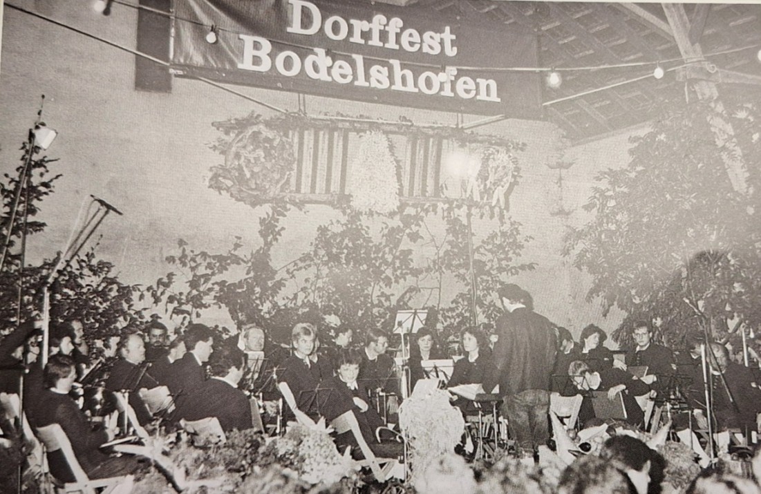 Schwarzweißaufnahme eines Blasorchesters mit Dirigenten unter einem Banner Dorffest Bodelshofen, geschmückt mit Zweigen und Lichterkette