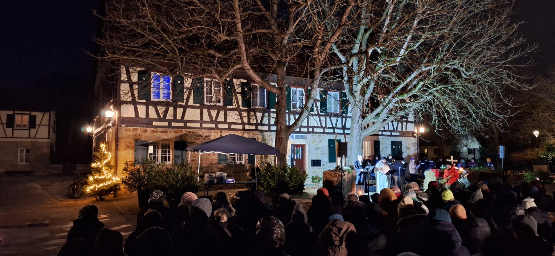 Abendliche Menschenmenge in Winterkleidung vor einem Fachwerkhaus, links ein hell erleuchteter Weihnachtsbaum, rechts Liturgen vor Strohballen und Holzkreuz