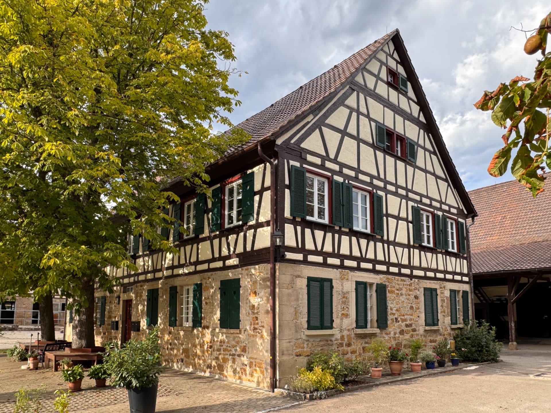Großes Fachwerkhaus mit Natursteinerdgeschoss, Topfpflanzen und belaubtem Baum vor gepflastertem Hof im Spätsommer