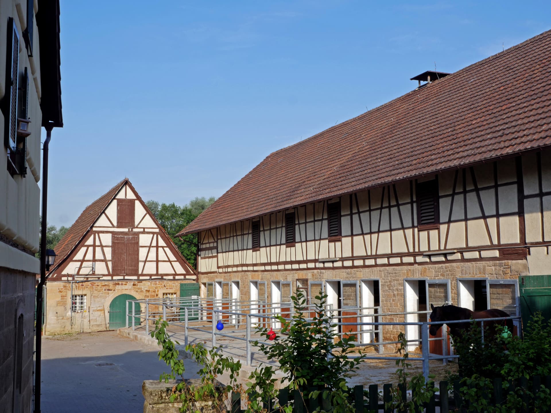 Fachwerk-Stallgebäude mit offenen Boxen, vorgelagerten Paddocks und einem dunkelbraunen Pferd